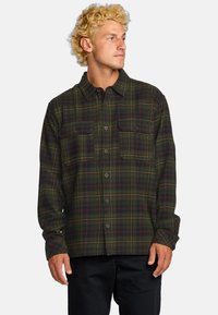 Camisa de franela a cuadros verdes con dos bolsillos en el pecho, cierre de botones y un corte holgado. La tela presenta líneas alternas oscuras y claras.