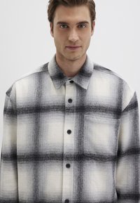 Checkered flannel shirt in black and white. Features a button-up front, folded collar, and a chest pocket with a soft texture.
