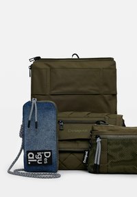Tre borse: uno zaino in nylon verde oliva con cerniere, una pochette in denim blu con una toppa logo, e un astuccio verde strutturato con cerniera.