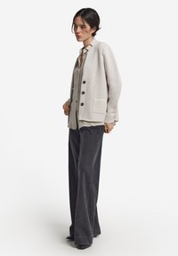 Jasno beżowy sweter z dzianiny z okrągłym dekoltem, długimi rękawami, z przodu na guziki i kieszenią. Połączony z szarym kołnierzykowym koszulą i ciemnymi szerokimi spodniami.