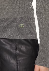 Gros plan sur une manche et un ourlet gris scintillant côtelés d'un pull avec un petit logo en métal, associés à un pantalon en cuir noir.