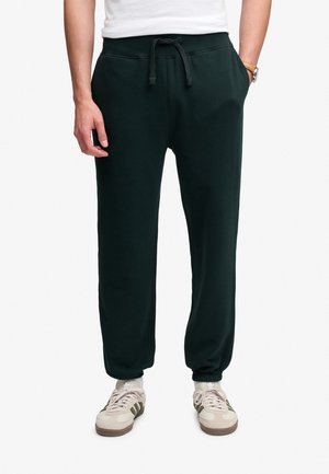 Man draagt donkergroene sweatpants met een koordje, witte sokken, beige sneakers met donkere strepen en een wit overhemd, staand met handen in de zakken.