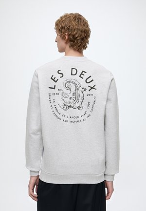 Personne aux cheveux courts et bouclés porte un sweat-shirt gris clair avec un graphisme noir et le texte "LES DEUX" ainsi qu'un dessin d'alligator dans le dos.