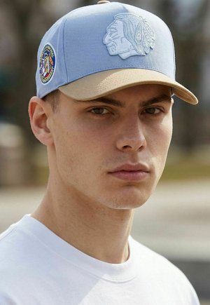 Junger Mann mit einer blau-beigen Baseballkappe mit einem bestickten Profil eines amerikanischen Ureinwohners und einem Aufnäher, gekleidet in einem weißen Hemd, draußen.