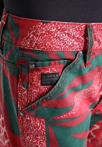 Rode en groene, met een patroon bedrukt broek van katoen, met een bloem- en geometrisch ontwerp, met een label waarop "G-STAR ORIGINALS RAW" staat.