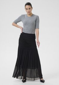 Grijs T-shirt met korte mouwen gecombineerd met een zwarte geplooide maxi-rok met ruches en een doorschijnende onderlaag. Zwarte puntige schoenen.