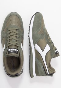 Zapatillas de color verde oliva con paneles de malla y detalles de ante. Presentan una marca en blanco y negro, puntera redondeada y suela de goma texturizada.