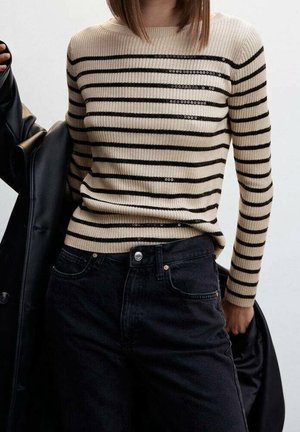 Personne portant un pull côtelé beige avec des rayures horizontales noires et un pantalon noir, tenant une veste noire sur une épaule.