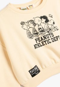 Sudadera color crema con un gráfico negro de personajes de Peanuts y el texto "PEANUTS ATHLETIC DEPT", con puños y dobladillo acanalados.