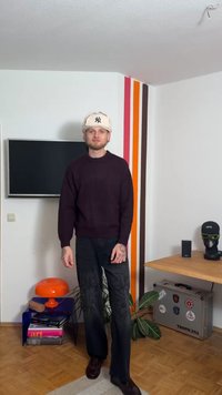 Purple knit sweater, black pants with decorative accents, beige cap, and dark shoes. Background includes a TV and a colorful striped wall.