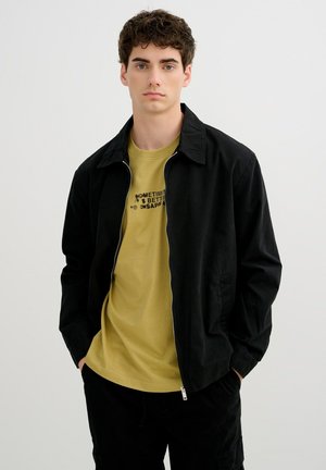 Joven con pelo corto y rizado que lleva una chaqueta negra con cremallera sobre una camiseta gráfica de color mostaza, de pie con las manos en los bolsillos.