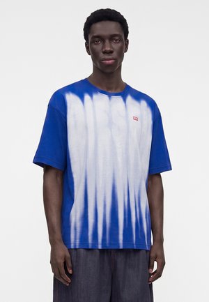 Jeune homme portant un t-shirt tie-dye Diesel bleu et blanc et un short sombre, debout devant un fond blanc uni.