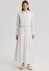 Robe blanche avec une jupe à volants et des accents en dentelle, associée à un gilet blanc ajusté et court avec des boutons. Complétée par des baskets beiges.
