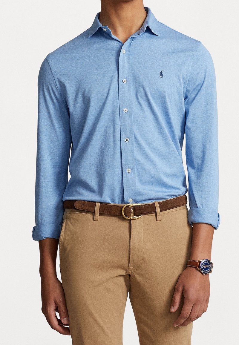 Jasnoniebieska koszula na długi rękaw z guzikami i kołnierzykiem, z małym logo na piersi, zestawiona z beżowymi spodniami i brązowym paskiem.