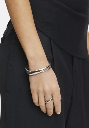 Zilveren metalen armband met een gedraaide uitstraling, soepel gepolijst. Een eenvoudige zilveren ring wordt ook op dezelfde hand gedragen, tegen donkere kleding.