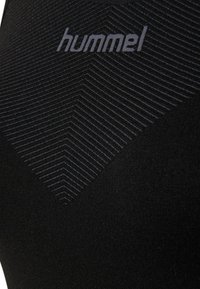 Musta urheilupaita, jossa on teksturoitu chevron-kuvio erottuvassa tummassa sävyssä, ja "hummel"-logo painettuna ylle. Sileä kangaspinta.