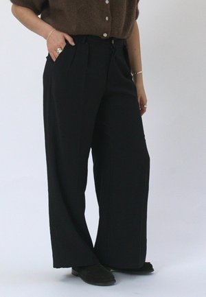 Persoon die zwarte, high-waist, wijd uitlopende broek draagt, een bruine, pluizige, met knoppen sluitende trui, zwarte schoenen, zilveren ringen en armbanden en met één hand in de zak staat.