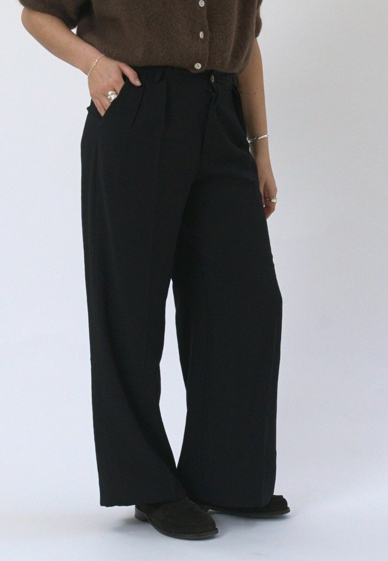Personne portant un pantalon noir taille haute à jambes larges, un pull marron duveteux boutonné, des chaussures noires et des bagues et bracelets en argent, debout avec une main dans la poche.