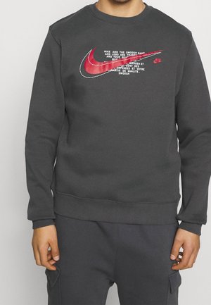 Sweat-shirt gris foncé avec un col rond, arborant un grand logo Swoosh Nike rouge et blanc ainsi qu'un design textuel à l'avant. Texture douce.