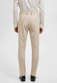 Bež hlače s gladko teksturo, zožan fit in zadnjimi žepi; oblečene z svetlo barvno majico z dolgimi rokavi. Črne čevlje.