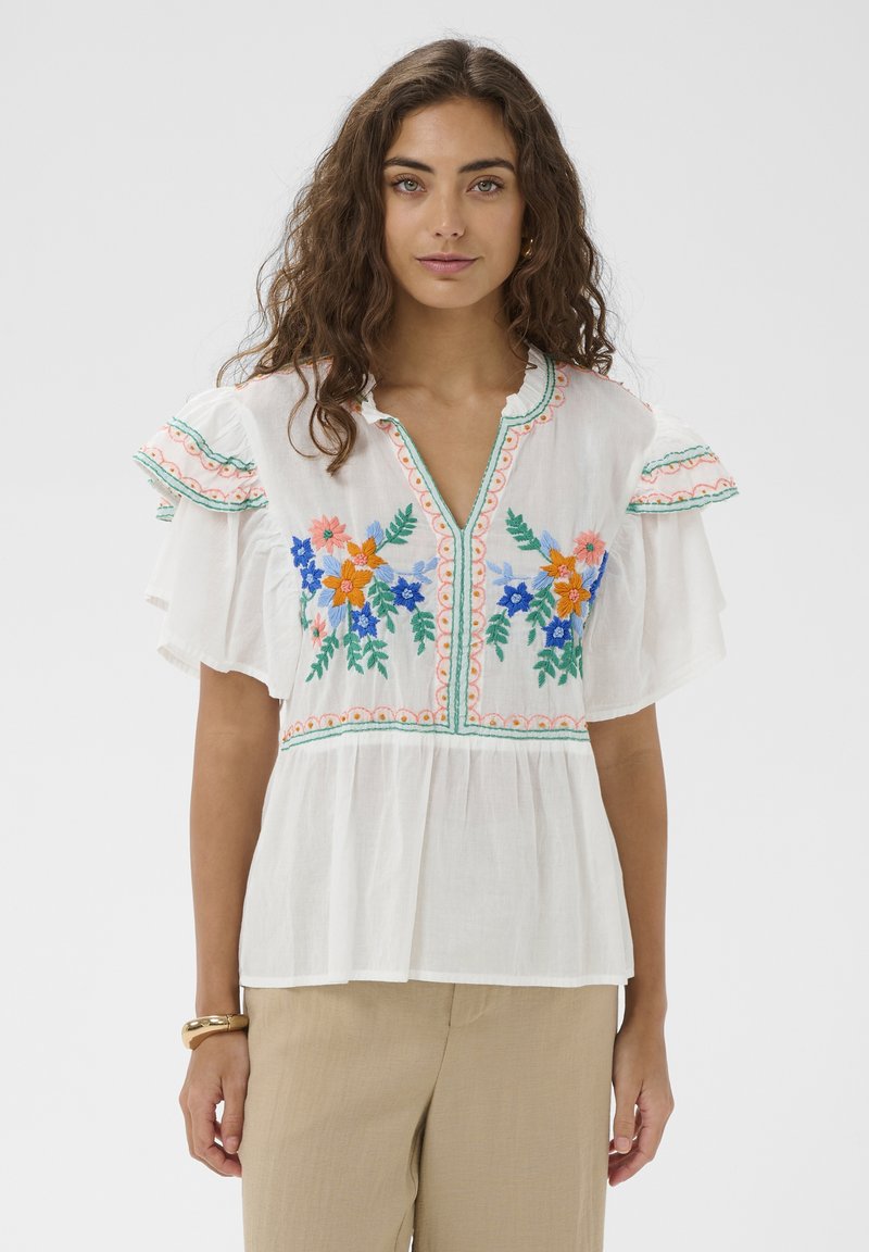 Femme portant un chemisier blanc avec une broderie florale colorée, des manches flottantes, et un pantalon beige, debout devant un fond clair uni.