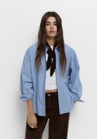 Camicia button-up azzurro chiaro con righe bianche verticali, abbinata a un top grigio e pantaloni in velluto a coste marroni. Una sciarpa scura è annodata attorno al collo.