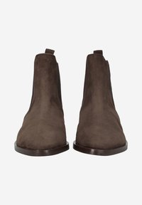 Bottines en suede marron avec un design épuré, dotées de panneaux latéraux élastiques et de languettes, reposant sur une semelle plate foncée.