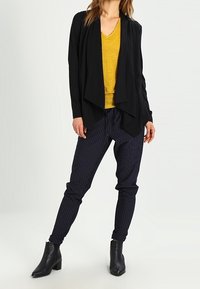 Svart draperad cardigan över en senapsgul v-ringad topp, tillsammans med marinblå randiga joggerbyxor och svarta ankelboots. Smidig textur och avslappnad passform.