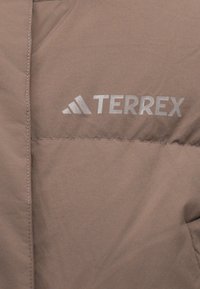 Pruun isoleeritud jope, millel on sile tekstuur, hõbedane "Terrex" logo ja nähtavad õmblused.