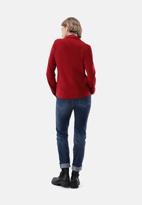Giacca rossa a maniche lunghe in un materiale morbido, abbinata a jeans in denim blu. Stivali neri alla caviglia con suola spessa completano l'outfit.