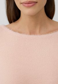 Pull en rose clair avec une texture douce et pelucheuse et un large décolleté. Le tissu semble léger et confortable, idéal pour une tenue décontractée.