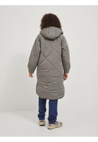 Abrigo largo gris acolchado con capucha, con patrones de diamantes y aberturas laterales, combinado con vaqueros azules y zapatillas beige.