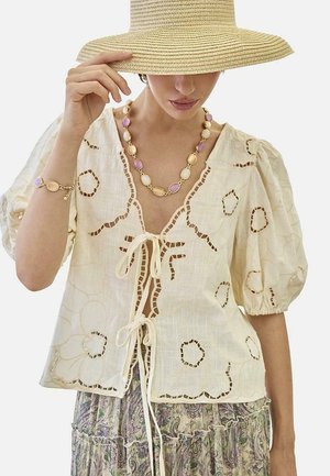 Mujer con un sombrero de paja de ala ancha, blusa crema bordada con lazos, collar de cuentas multicolores, pulsera y falda estampada.