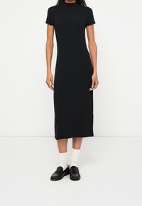 Personne portant une robe midi noire à manches courtes, des chaussettes blanches et des mocassins noirs, debout devant un fond blanc uni.
