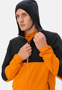 Giacca a zip arancione e nera con cappuccio, polsini elastici e design a blocchi di colore. Tessuto leggero con una texture liscia.