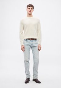 Gaiši bēša adīta džemperis, gaiši zili džinsa bikses, brūna ādas josta, tumši brūni zābaki, stāvoša poza uz vienkārša balta fona.