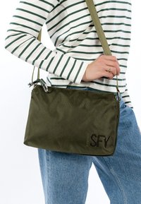 Zielona teksturowana torba na ramię o prostokątnym kształcie, z logo, regulowanym paskiem i srebrnymi ozdobami.