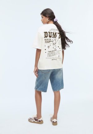 T-shirt oversized blanche avec impression et graphiques noirs au dos, assorti à un short en jean et des baskets marron à motifs.