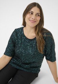 Femeie cu păr lung, castaniu, îmbrăcată cu un top scurt cu mâneci, de culoare verde închis, cu paiete, și pantaloni negri, stând și zâmbind, privindu-se pe o parte.