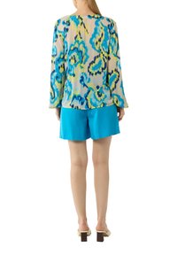 Blumenmuster langärmlige Bluse in Blau, Grün und Gelb auf einem hellen Hintergrund, kombiniert mit knallblauen Shorts und cremefarbenen Blockabsatzschuhen.