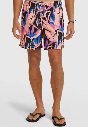 Man met roze, paarse, blauwe en zwarte zwemshorts met een tropische print, een perzikkleurige trekkoord en zwarte slippers, op een witte achtergrond.