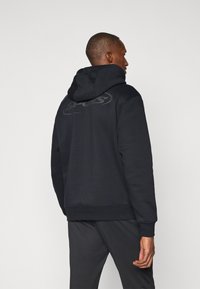Black hoodie with a kangaroo pocket, ribbed cuffs and hem, featuring a tonal logo on the back. Material appears soft and textured.