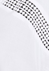 Tissu blanc avec un motif texturé noir sur l'épaule, présentant de petits cercles et des accents de design géométrique. Texture globale lisse.