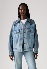 Veste en denim bleu clair au design classique, avec un empiècement en pointe, deux poches poitrine et des boutons marron. Tissu texturé, coupe décontractée.