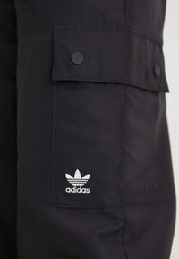 Mustad mustad püksid, mis on valmistatud vastupidavast kangast, kahe nupuga taskuga ja vasakul reitel valge broderi Adidas logo.