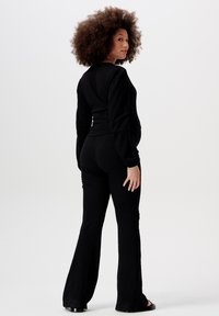 Schwarzes Langarmshirt mit strukturiertem Stoff, kombiniert mit eng anliegenden schwarzen Schlaghosen. Das Modell steht seitlich vor einem neutralen Hintergrund.