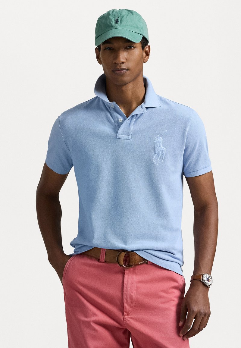 Light blue polo shirt with a textured fabric and embroidered logo, paired with pink shorts and a brown belt. Green cap and watch visible.