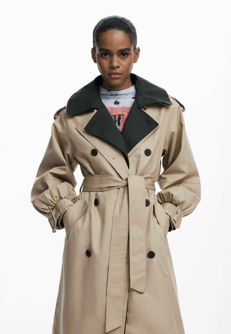 Manteau beige avec un col noir, design à double boutonnage, manches cintrées et taille nouée, comportant deux poches latérales et des boutons noirs.