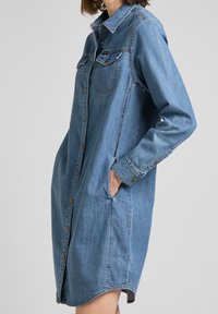 Jeansklänning i ljusblå med långa ärmar, knappstängning fram, bröstfickor och sidfickor. Krum form på fållen och mjuk textur.