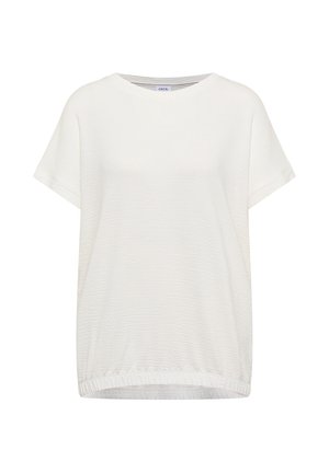 Kvinde smilende, går indendørs iført en hvid kortærmet top, løse grønne bukser og lyse sneakers med grønne detaljer.